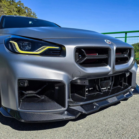 A sleek car with aggressive headlights and a carbon fiber front bumper is parked on asphalt, under a clear blue sky, with green railing and trees in the background.