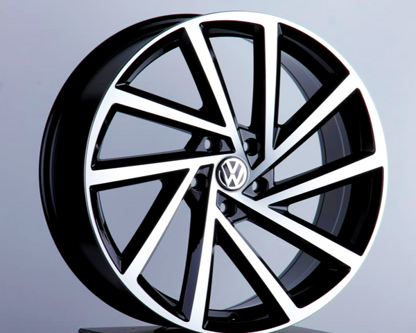 A stylish black and silver alloy wheel, featuring the Volkswagen logo, is displayed upright on a dark stand against a plain gray background.