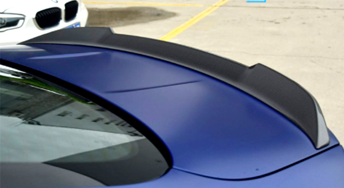 A black carbon fiber spoiler sits on the rear of a blue car, with a white car parked nearby in an outdoor concrete area marked by yellow lines.