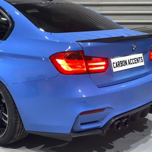 A blue car with illuminated taillights is parked in an indoor garage. The license plate reads, "CARBON ACCENTS." The car has dual exhausts and a carbon-fiber rear spoiler.