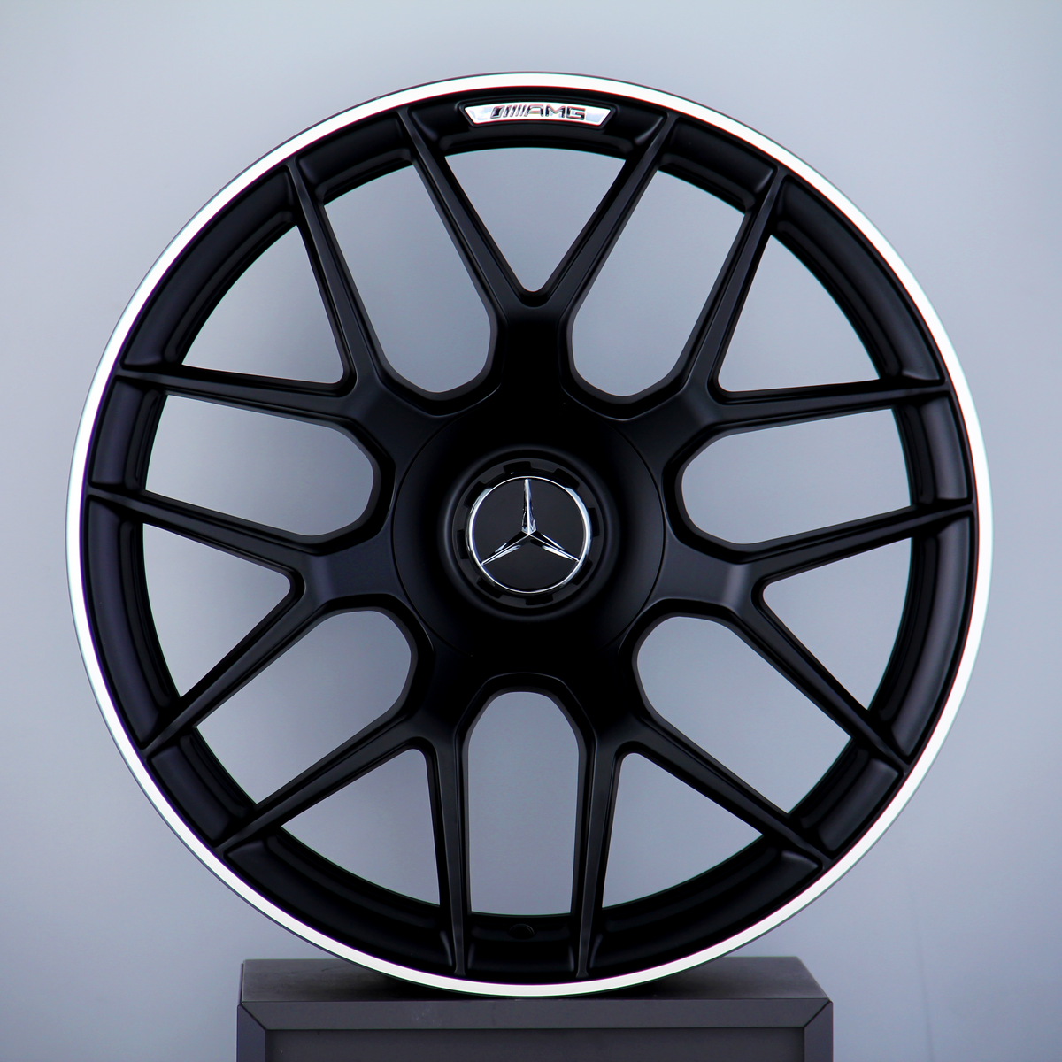 A black multi-spoke car wheel rim with a Mercedes-Benz logo at the center, labeled "AMG," is displayed against a neutral background, resting atop a pedestal.