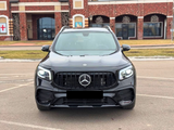 A black Mercedes SUV is parked, facing forward, in an outdoor lot with a brick building backdrop. The car's lights are on, and it features distinctive grille and emblem design.