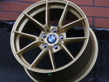 A gold BMW wheel rim rests against a brick-patterned wall on asphalt. The center features the BMW logo with spokes radiating outward.