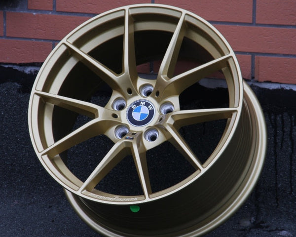 A gold BMW wheel rim rests against a brick-patterned wall on asphalt. The center features the BMW logo with spokes radiating outward.