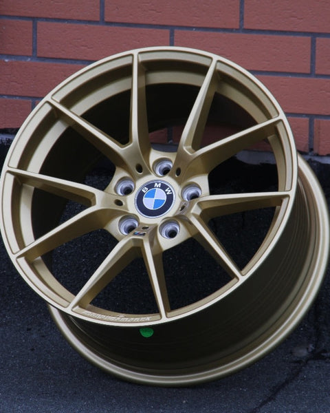 A gold BMW wheel rim rests against a brick-patterned wall on asphalt. The center features the BMW logo with spokes radiating outward.