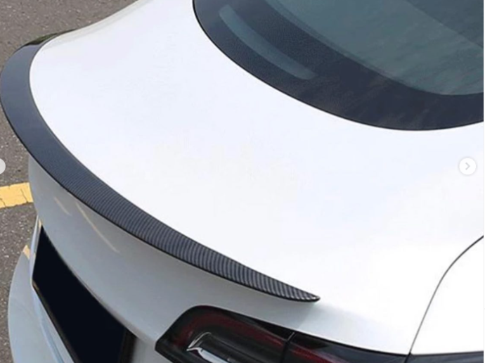 A white car features a carbon-fiber rear spoiler, parked on a paved road with yellow line markings, viewed from the rear side, capturing the car's sleek design.