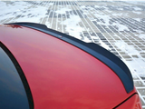 A red car's rear spoiler sits prominently against a snowy, grid-patterned ground. A small emblem or badge is affixed to the spoiler's center.