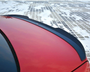 A red car's rear spoiler sits prominently against a snowy, grid-patterned ground. A small emblem or badge is affixed to the spoiler's center.