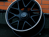 A black AMG car wheel rests against an orange brick wall. The wheel features a Mercedes logo at its center.
