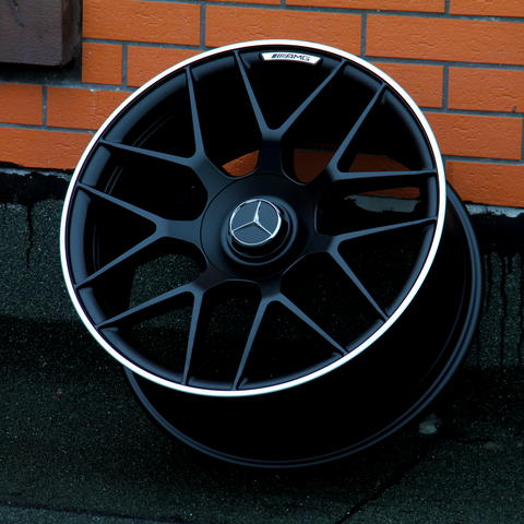 A black AMG car wheel rests against an orange brick wall. The wheel features a Mercedes logo at its center.