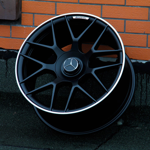 A black alloy wheel with a Mercedes emblem and "AMG" text is propped against an orange brick wall. The ground appears to be a dark, textured surface.