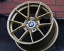 A gold BMW alloy wheel rests against a brick wall on an asphalt surface. The BMW logo is centered on the wheel's hub.