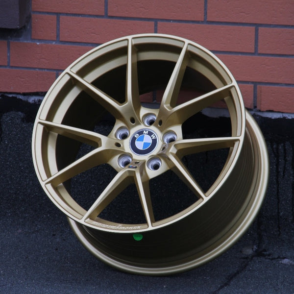 A gold BMW alloy wheel rests against a brick wall on an asphalt surface. The BMW logo is centered on the wheel's hub.
