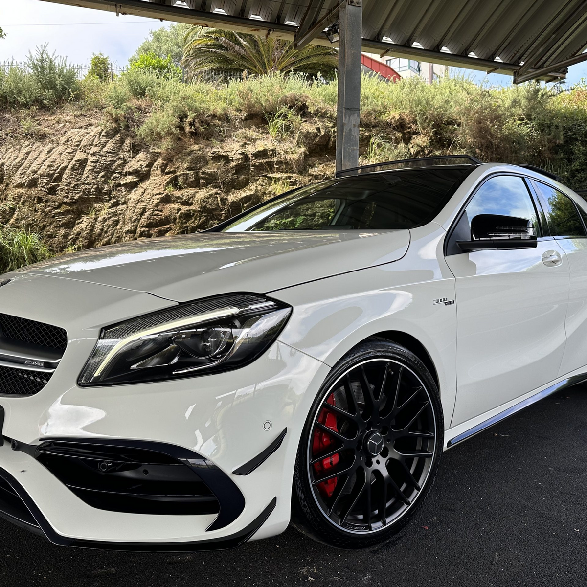 A white Mercedes-Benz sedan is parked under a metal-roofed structure, featuring sleek black wheels and set against a backdrop of rocks and dense shrubs.
