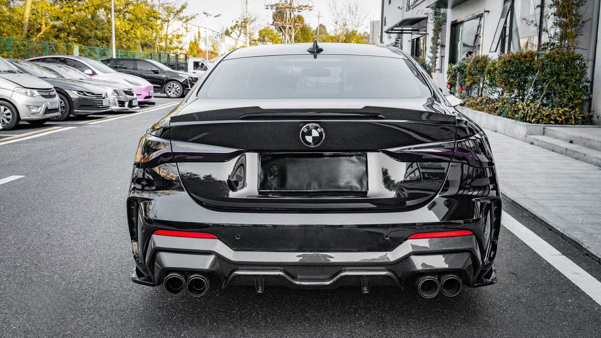 A black BMW car parked on a street, viewed from behind, surrounded by trees and other parked vehicles. The environment is urban, with buildings visible on the right.
