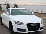 A white Audi car, parked on a paved road, displays a prominent logo on the front grille. The background features a white fence and sparse trees against a cloudy sky.