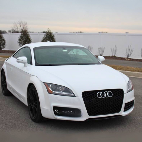 A white Audi car, parked on a paved road, displays a prominent logo on the front grille. The background features a white fence and sparse trees against a cloudy sky.