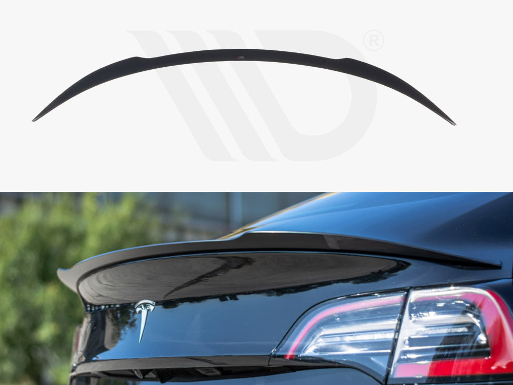 A black car spoiler is attached to the rear of a blue vehicle with a visible trunk emblem. The background is blurred, featuring greenery and a building.