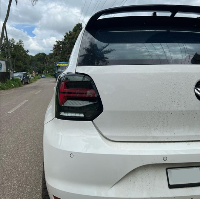 A white car is parked on a roadside, showcasing its rear end with a visible taillight. Surrounding it are trees, a blue vehicle, and a partly cloudy sky.
