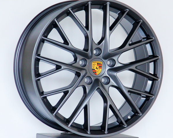 A dark metallic alloy wheel with multiple spokes and a central emblem featuring the Porsche logo, displayed on a platform against a plain white background.