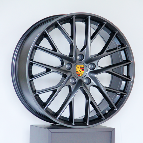 A dark metallic alloy wheel with multiple spokes and a central emblem featuring the Porsche logo, displayed on a platform against a plain white background.