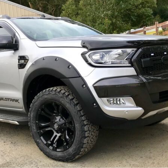 A silver pickup truck with "WILDTRAK" on the side is parked on a gravel driveway beside a building, surrounded by greenery. The front grille displays the "FORD" logo prominently.