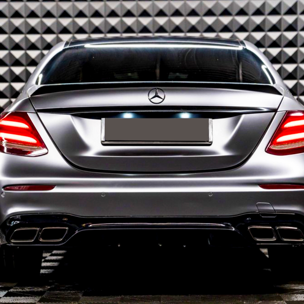 A sleek car with illuminated taillights is parked, facing a geometric-patterned wall in a showroom-like setting.
