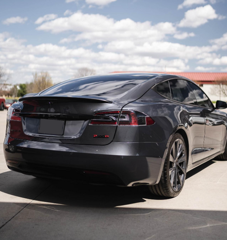 A sleek grey car is parked on a concrete surface under a partly cloudy sky. The vehicle's design and polished finish are highlighted. The rear badge reads "P100D".