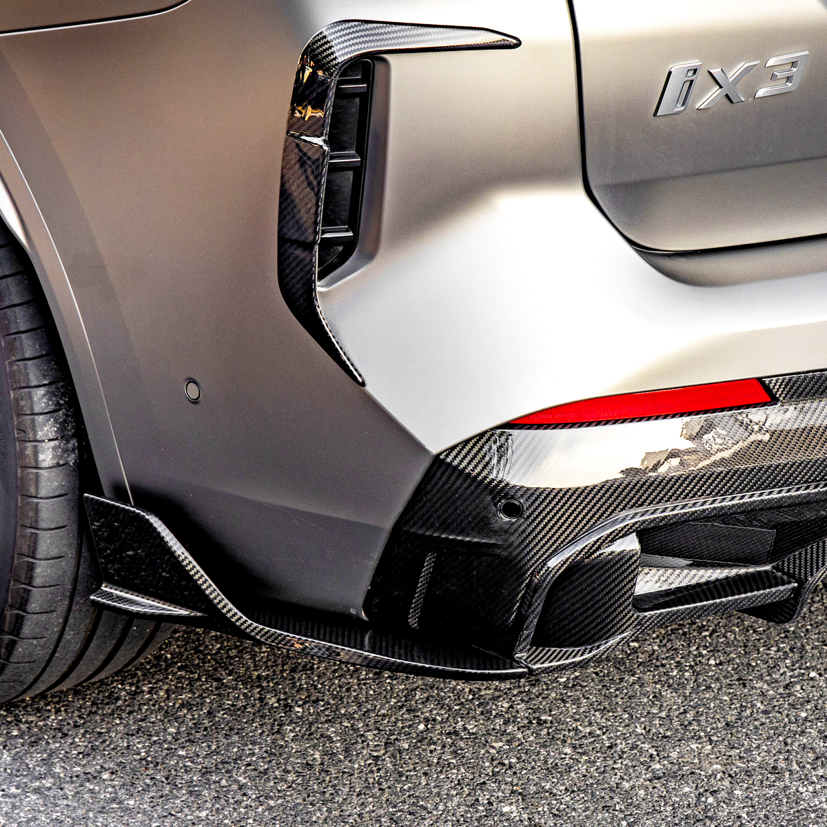 Car with "iX3" badging, featuring a sleek rear bumper and carbon fiber diffuser, parked on a paved surface. The rear wheel has a modern rim design.