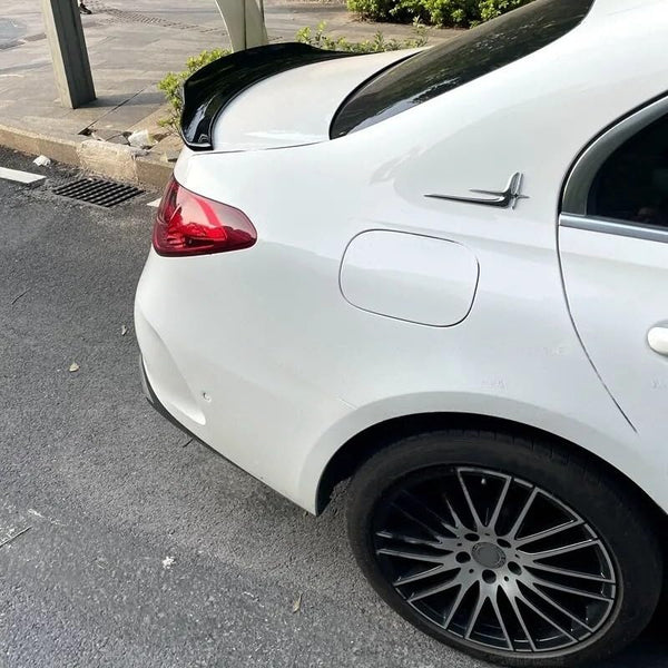 White car's rear section with a black spoiler, parked on a gray asphalt road. Nearby, there are plants and a sidewalk with pavement.