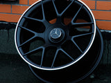 A black Mercedes-Benz AMG wheel rests against a brick wall on a dark, textured ground. The wheel features a silver trim and the Mercedes-Benz logo at the center.