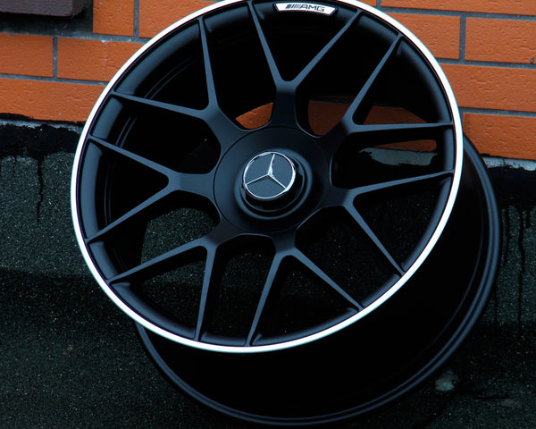 A black Mercedes-Benz AMG wheel rests against a brick wall on a dark, textured ground. The wheel features a silver trim and the Mercedes-Benz logo at the center.