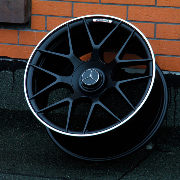 A black Mercedes-Benz AMG wheel rests against a brick wall on a dark, textured ground. The wheel features a silver trim and the Mercedes-Benz logo at the center.