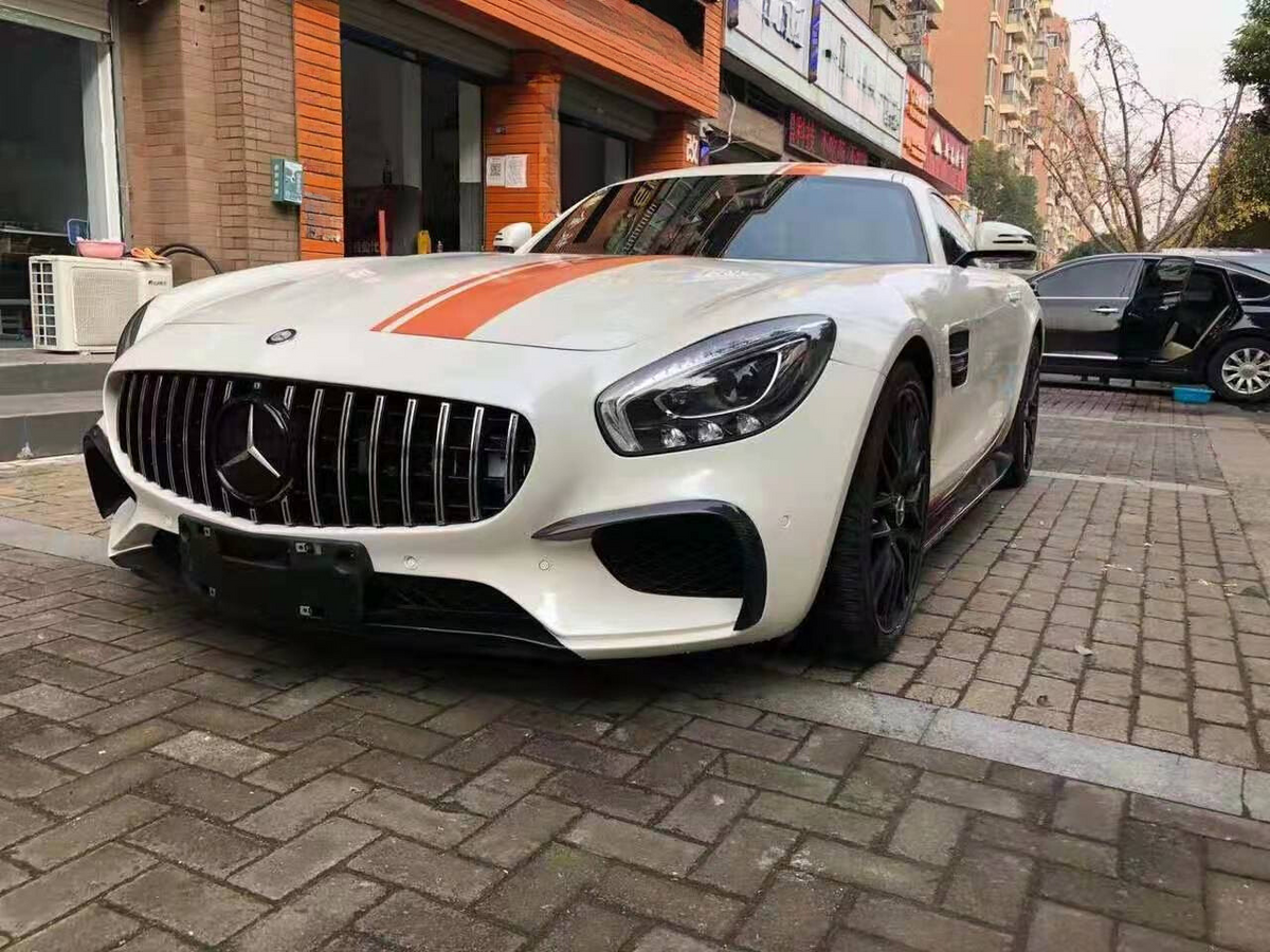 A white sports car with an orange stripe is parked on a brick-paved street. Nearby, a building with signs and another car with an open door are visible.