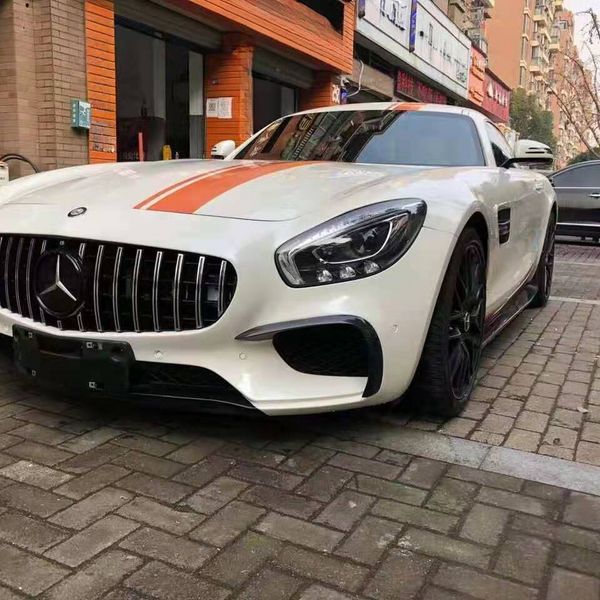 A white sports car with an orange stripe is parked on a brick-paved street. Nearby, a building with signs and another car with an open door are visible.