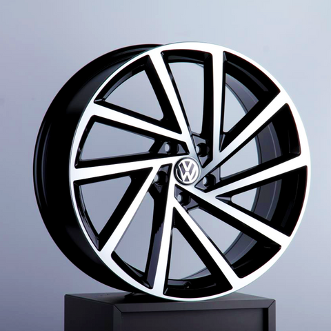 A black and silver alloy car wheel, featuring a Volkswagen logo at the center, is displayed upright on a pedestal against a plain, light gray background.