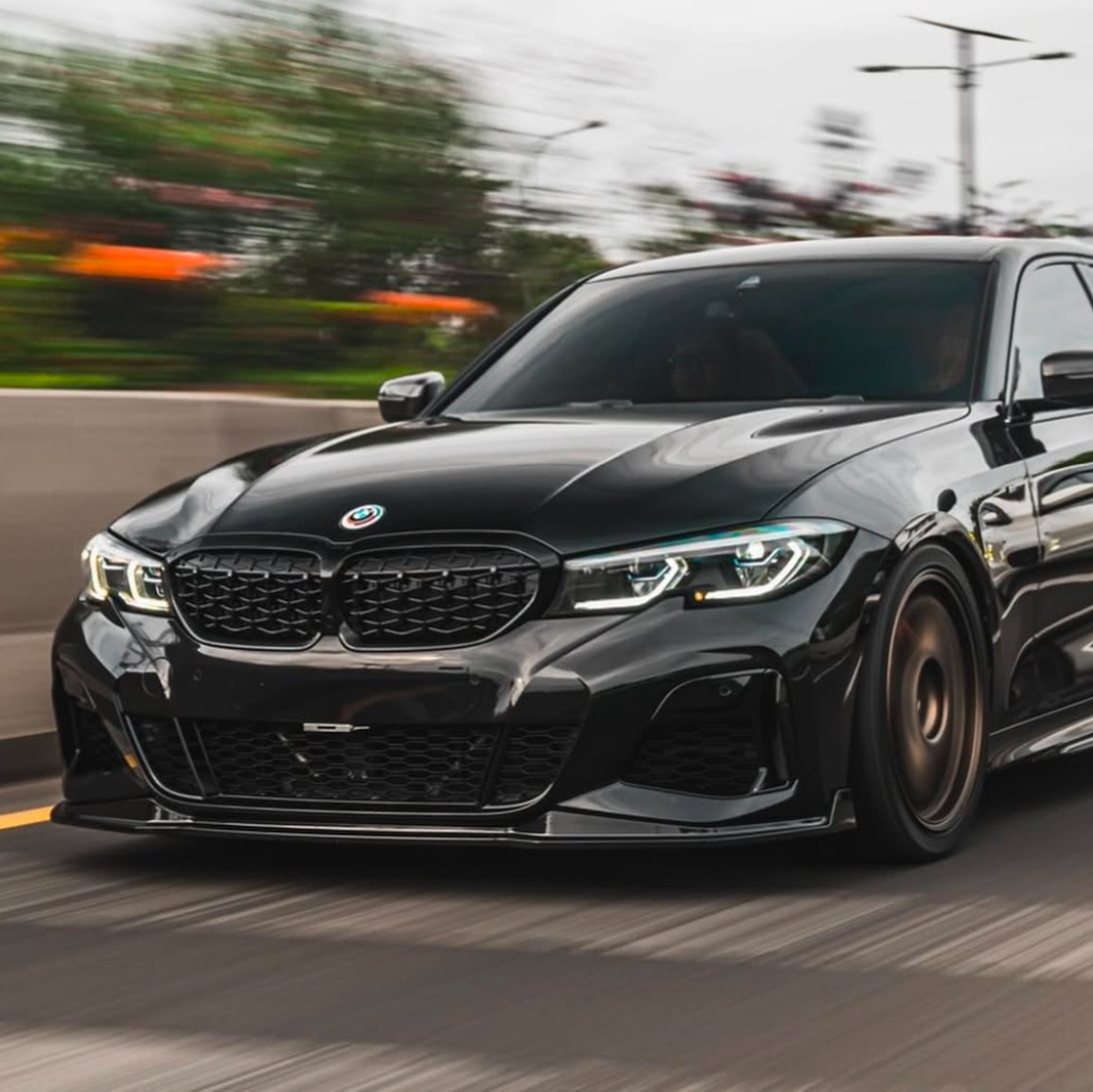 A black car with sleek design speeds along a highway, surrounded by blurred greenery and streetlights, highlighting motion. The BMW logo is visible on the front grille.