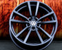 A black, multi-spoke Volkswagen wheel rim rests against an orange, textured background on a gray surface. The Volkswagen logo is prominently displayed in the center.