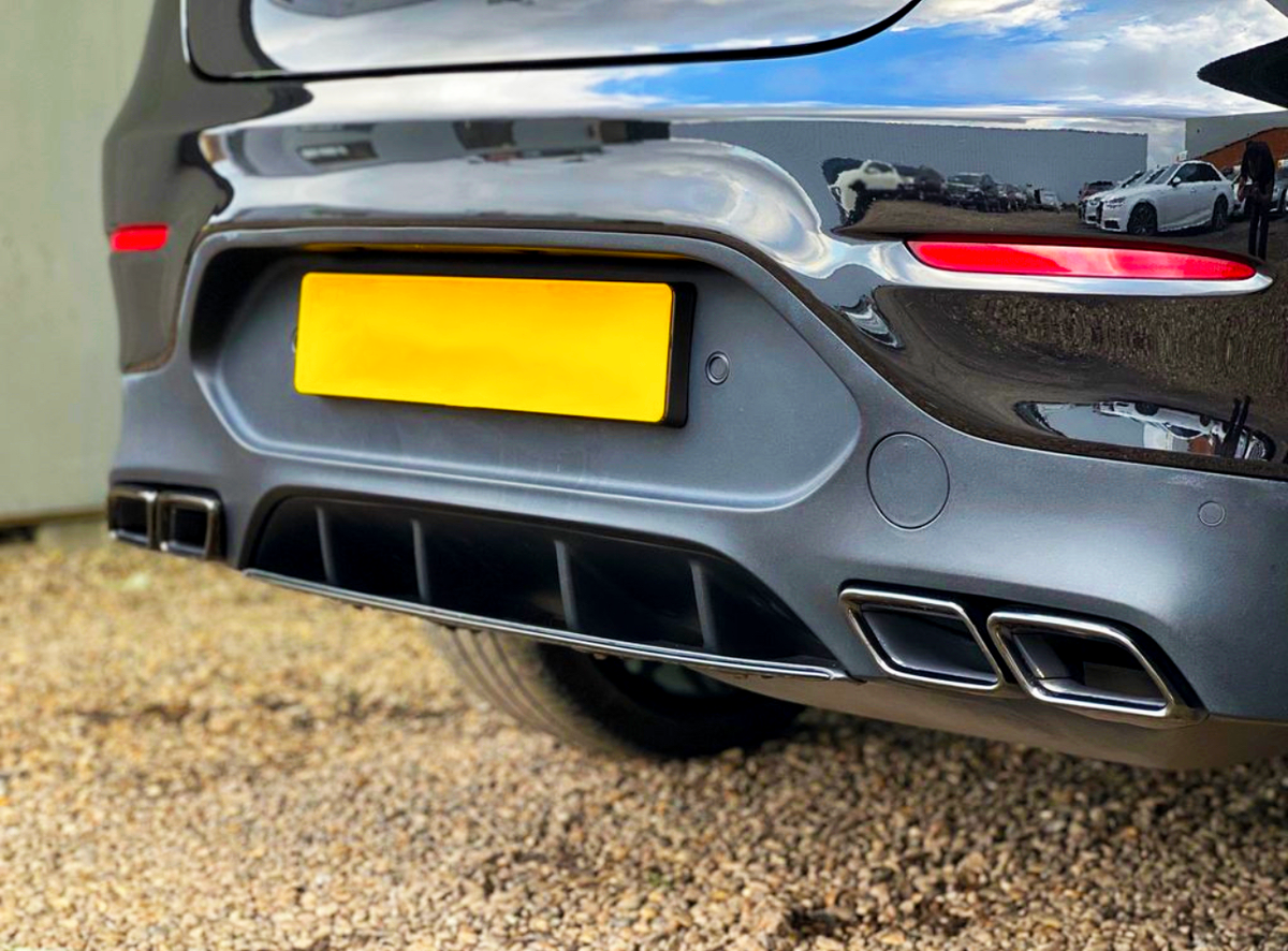 The rear end of a dark car is parked on gravel, featuring a bright yellow license plate, dual exhausts, and red reflectors. Nearby, other vehicles are visible in the reflection.