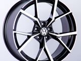 A black and silver alloy wheel with a Volkswagen logo in the center is positioned upright against a plain white background, highlighting its intricate spoke design.