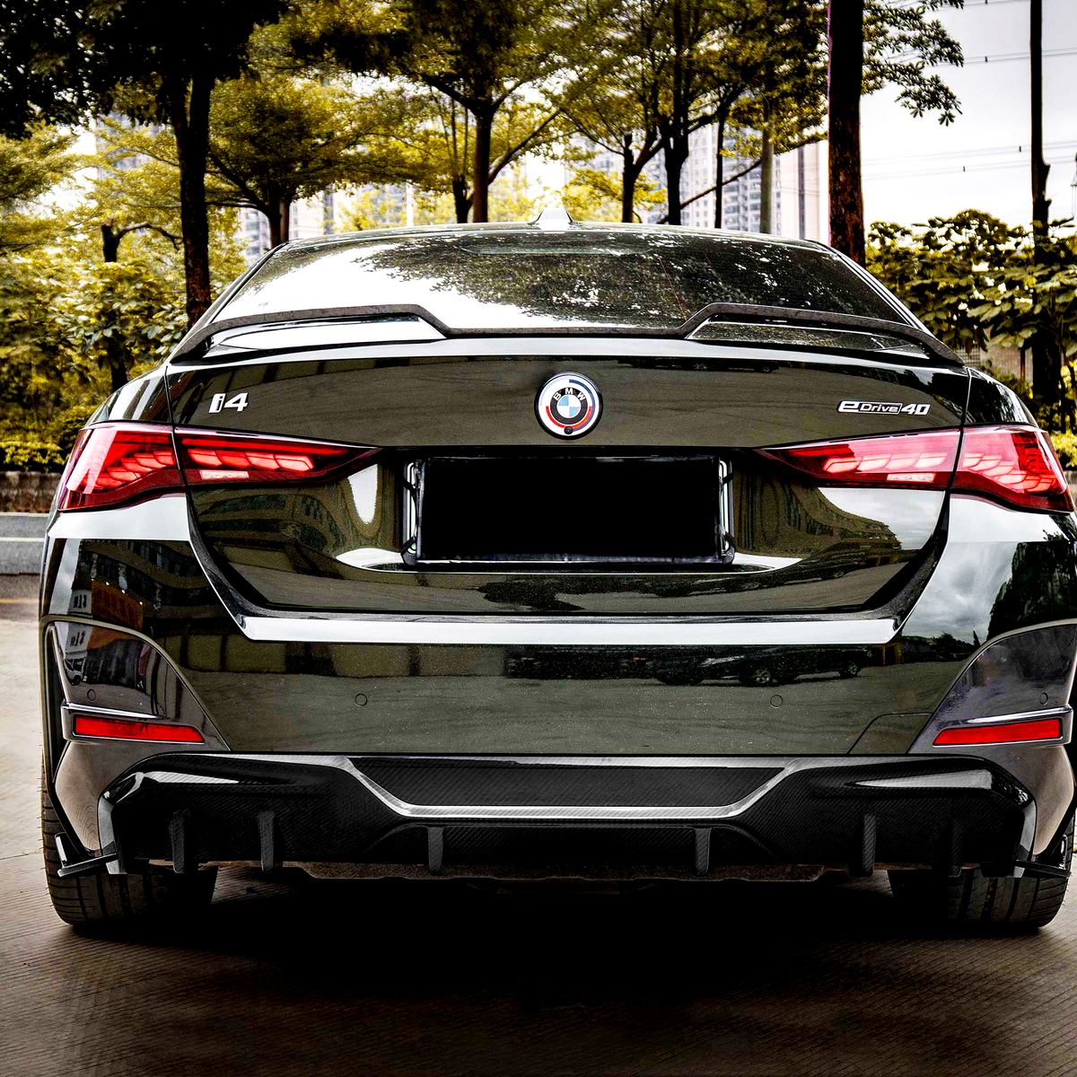 A sleek black car is parked, displaying a rear view with red taillights illuminated. The environment features green trees and a city street. Visible text includes: "i4" and "eDrive40."