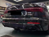 A sleek black Audi A6L with dual exhausts is parked indoors under bright, square ceiling lights. Wall art and a shelf with trophies are visible. Text: 