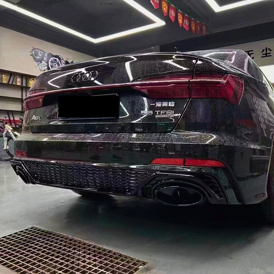 A sleek black Audi A6L with dual exhausts is parked indoors under bright, square ceiling lights. Wall art and a shelf with trophies are visible. Text: "A6L", "55 TFSI".