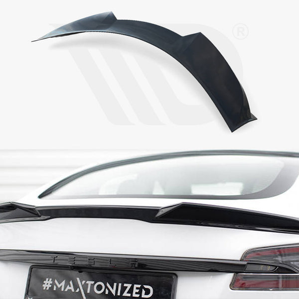 A black car spoiler is positioned on the rear of a white vehicle, with the text "#MAXTONIZED" on the license plate. It rests in a bright, indoor environment.