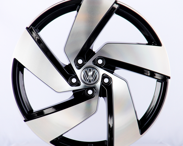 A metallic car wheel rim with a five-spoke design features a central logo containing a "VW" emblem, displayed against a plain white background.