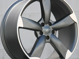 A metallic car wheel rim with five wide spokes is positioned upright. It's placed on a reflective surface in front of a neutral, light-colored wall.