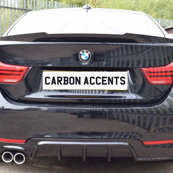 Car with dual exhaust displays "CARBON ACCENTS" on the license plate; parked on a paved surface with metal fencing and greenery in the background.