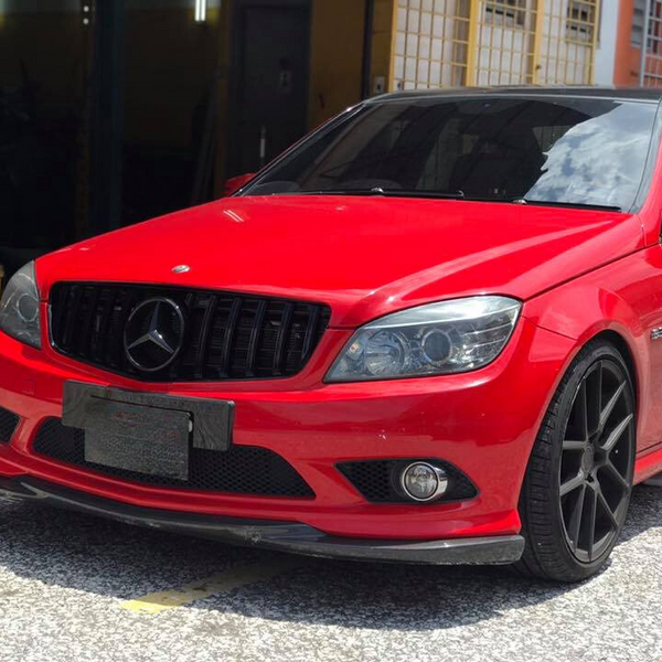 Red Mercedes-Benz car parked on a sunlit street, featuring black rims and tinted windows. Nearby, there are buildings with yellow grids and a partially open garage door.