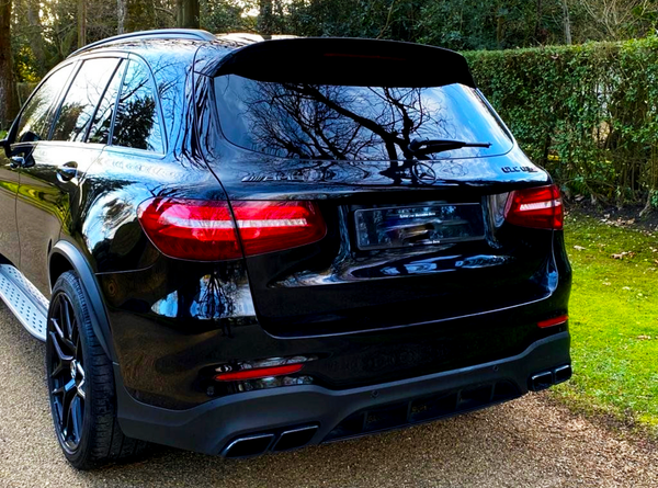 A black SUV is parked on a gravel path, surrounded by trees and hedges. Reflections of branches appear on its windows. The text on the car reads "GLC 43" and "AMG".