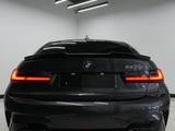 A dark-colored BMW 330i is parked indoors, showing its illuminated rear lights. The sleek, modern ceiling features hanging fluorescent lights, adding a contemporary ambiance to the garage setting.